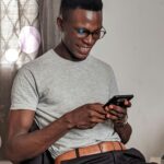 A happy young man uses his smartphone while sitting indoors, expressing joy.